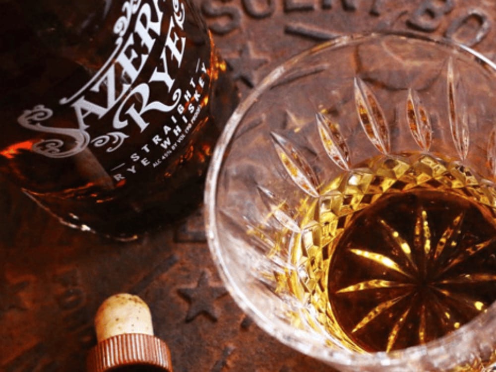 Bottle of Sazerac whiskey with a glass filled with whiskey on a rustic surface. Available for purchase at Blenheim Vintners.